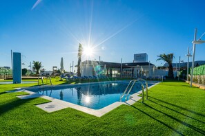 A swimming pool with a sunlit sky, a building with a sign, and green grass. - Camping Sol de Calpe Boreal - Caravan park (Calpe)