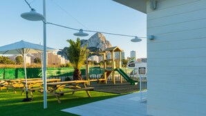 A playground with picnic tables and a climbing structure, set against a backdrop of buildings and a mountain. - Camping Sol de Calpe Boreal - Caravan park (Calpe)