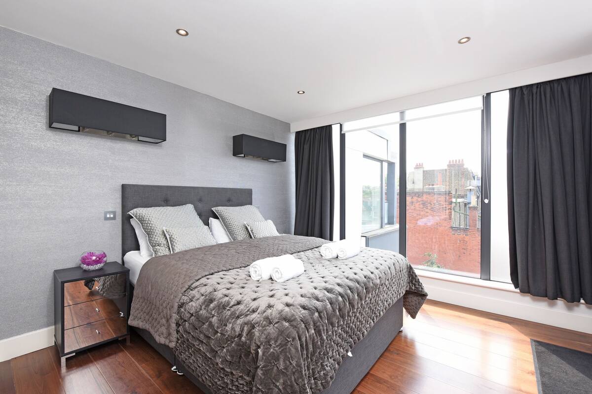 A modern bedroom with a large bed, a nightstand, and a view of the city through large windows.