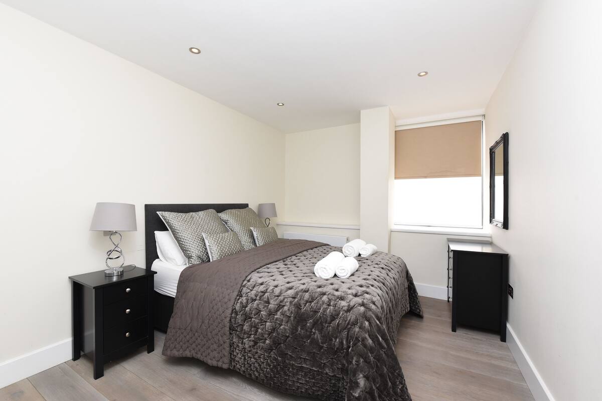 A bedroom with a bed, a nightstand, a lamp, a window with a blind, and a mirror on the wall.