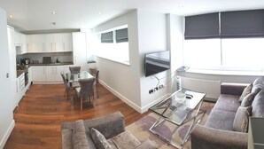 Un appartement moderne comprenant une cuisine, un coin repas et un salon. Il est équipé d’un téléviseur à écran plat, d’une table basse en verre et d’un canapé.