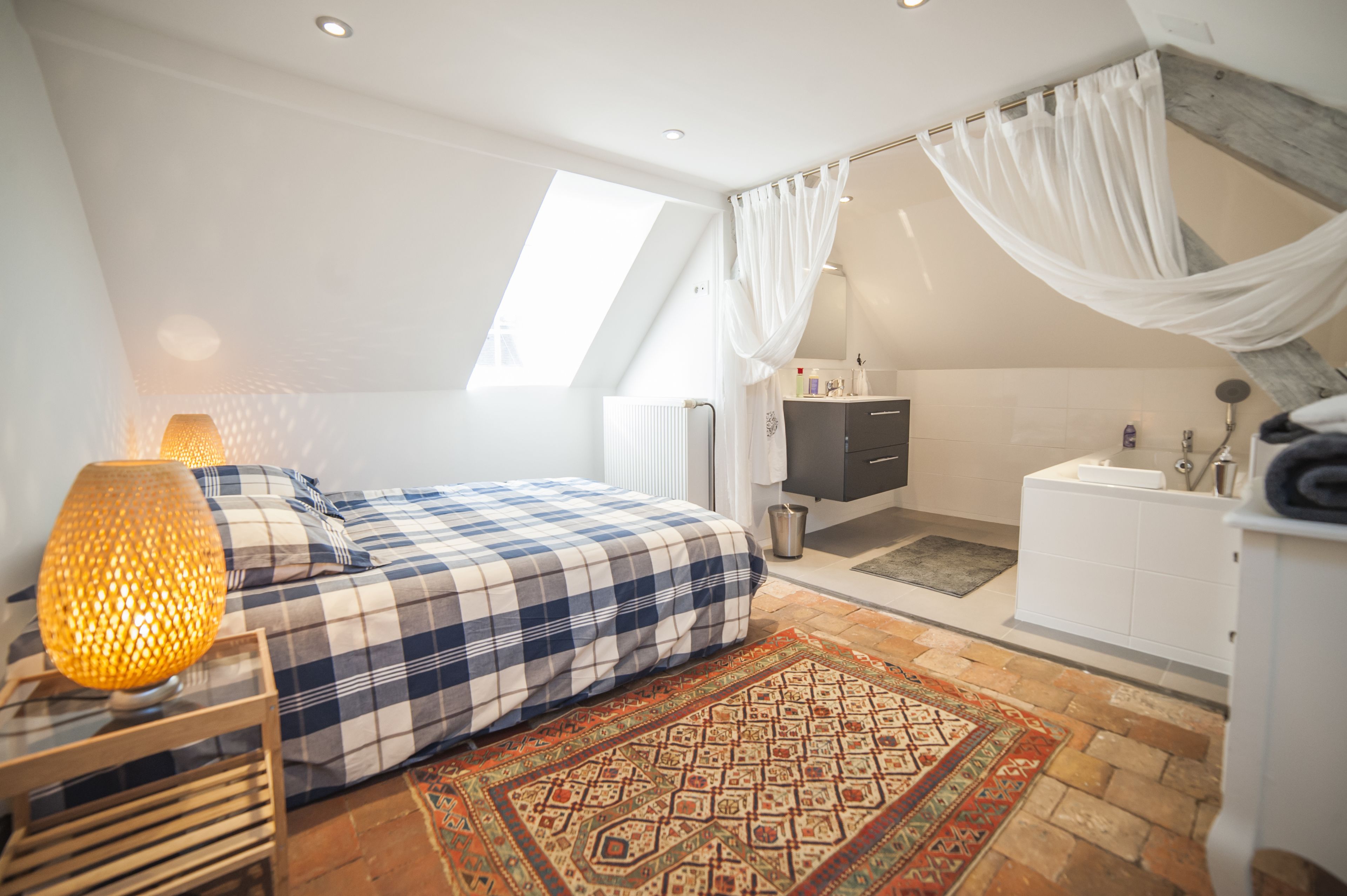 A bedroom with a bed, a nightstand, a lamp, a window, a bathroom with a shower curtain, and a rug.