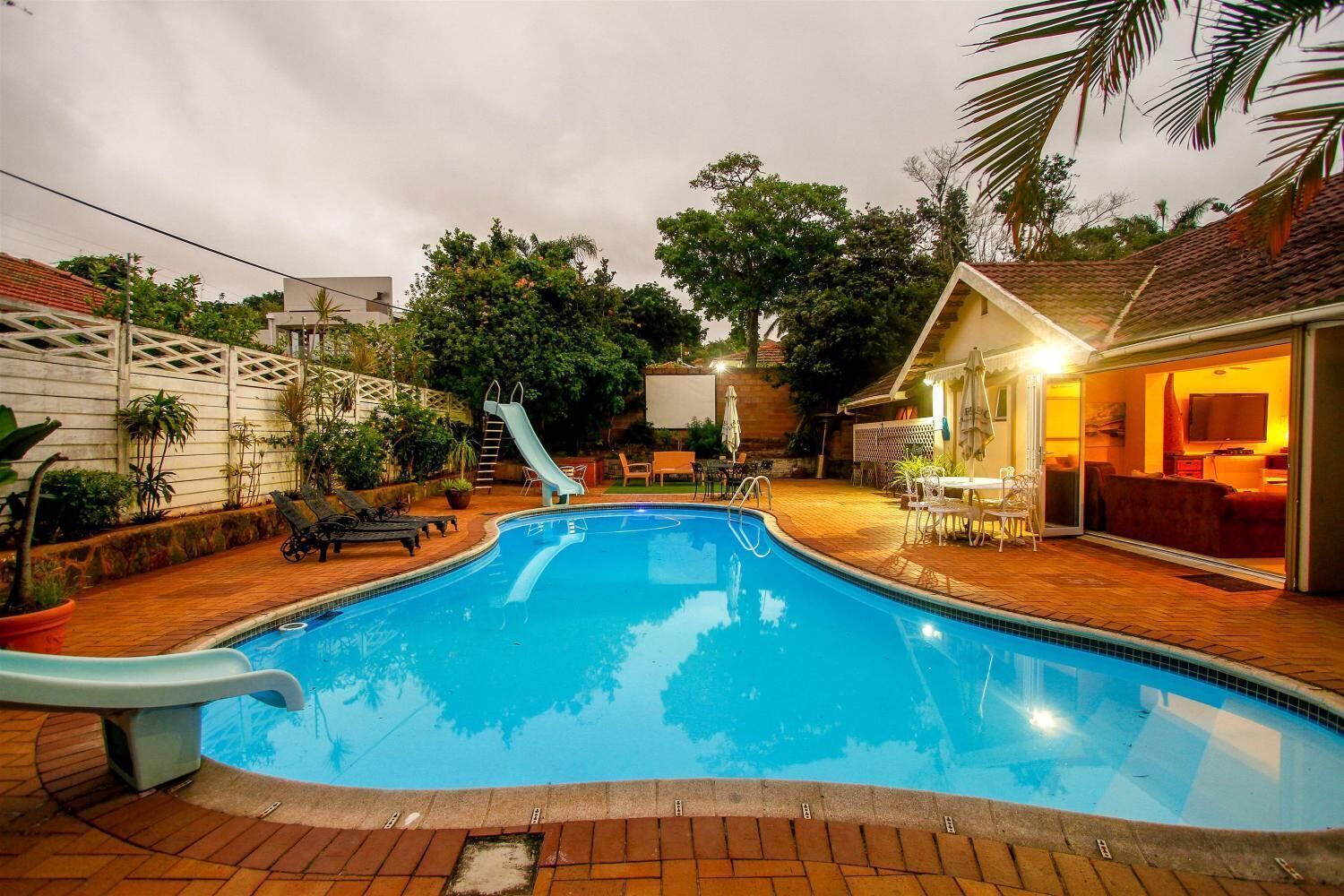 A swimming pool with a slide, surrounded by a patio area with outdoor furniture and a small building with a TV.
