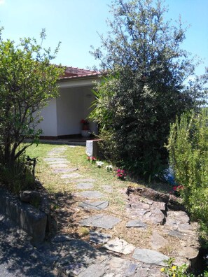 A stone pathway leading to a building with a red roof, surrounded by greenery and flowering plants.