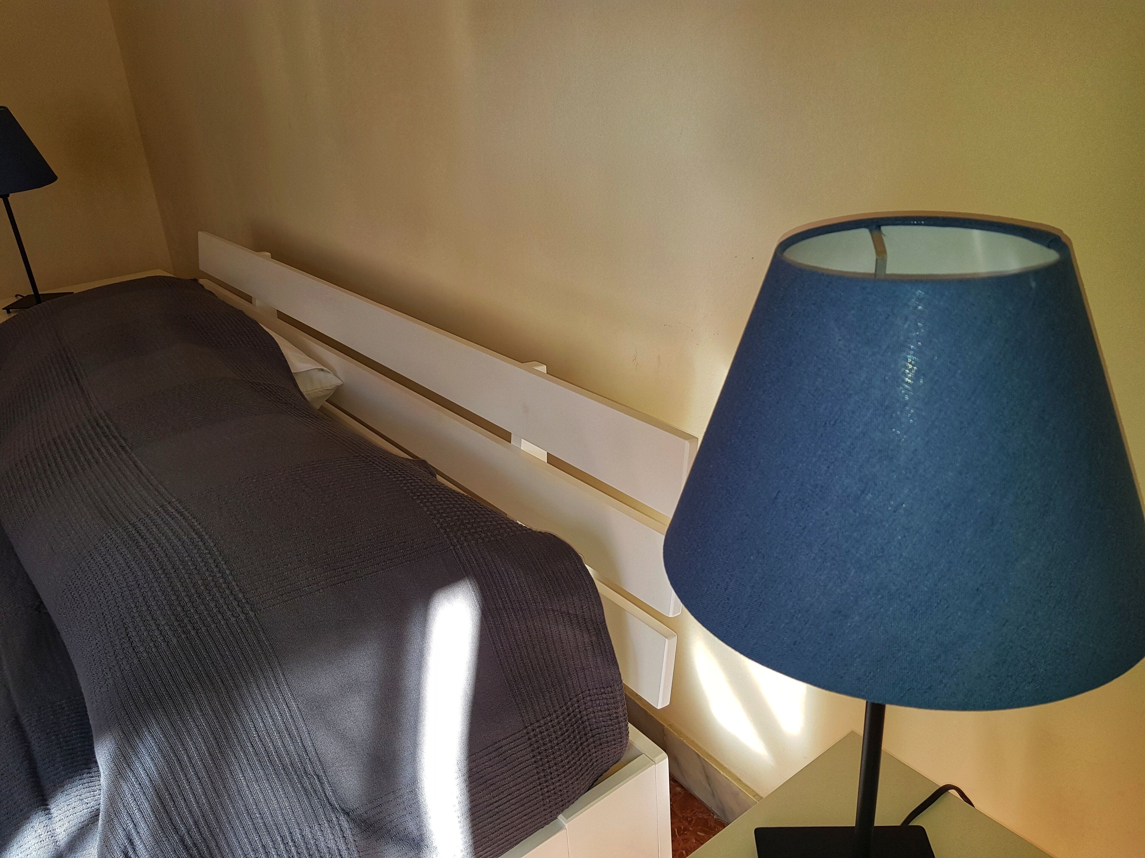 Ein Bett mit dunkler Decke und einer blauen Lampe daneben.