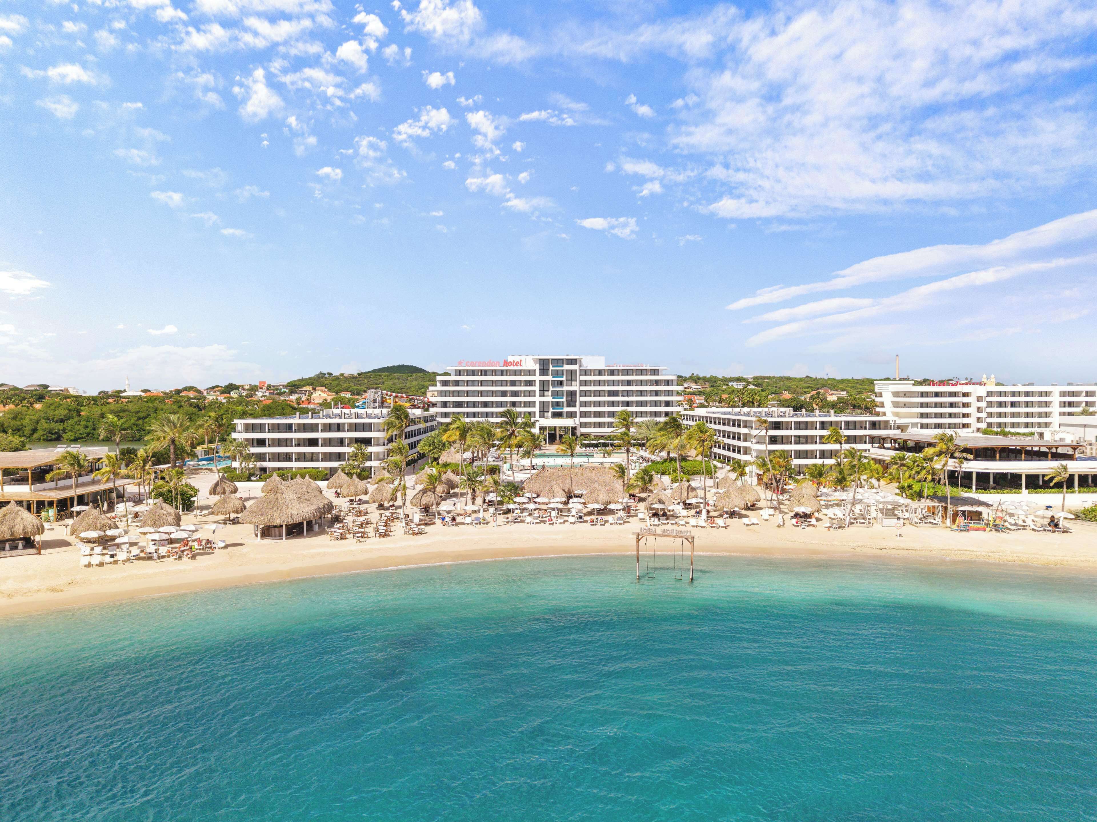 A coastal hotel with multiple buildings, a beach with umbrellas, and clear blue water.