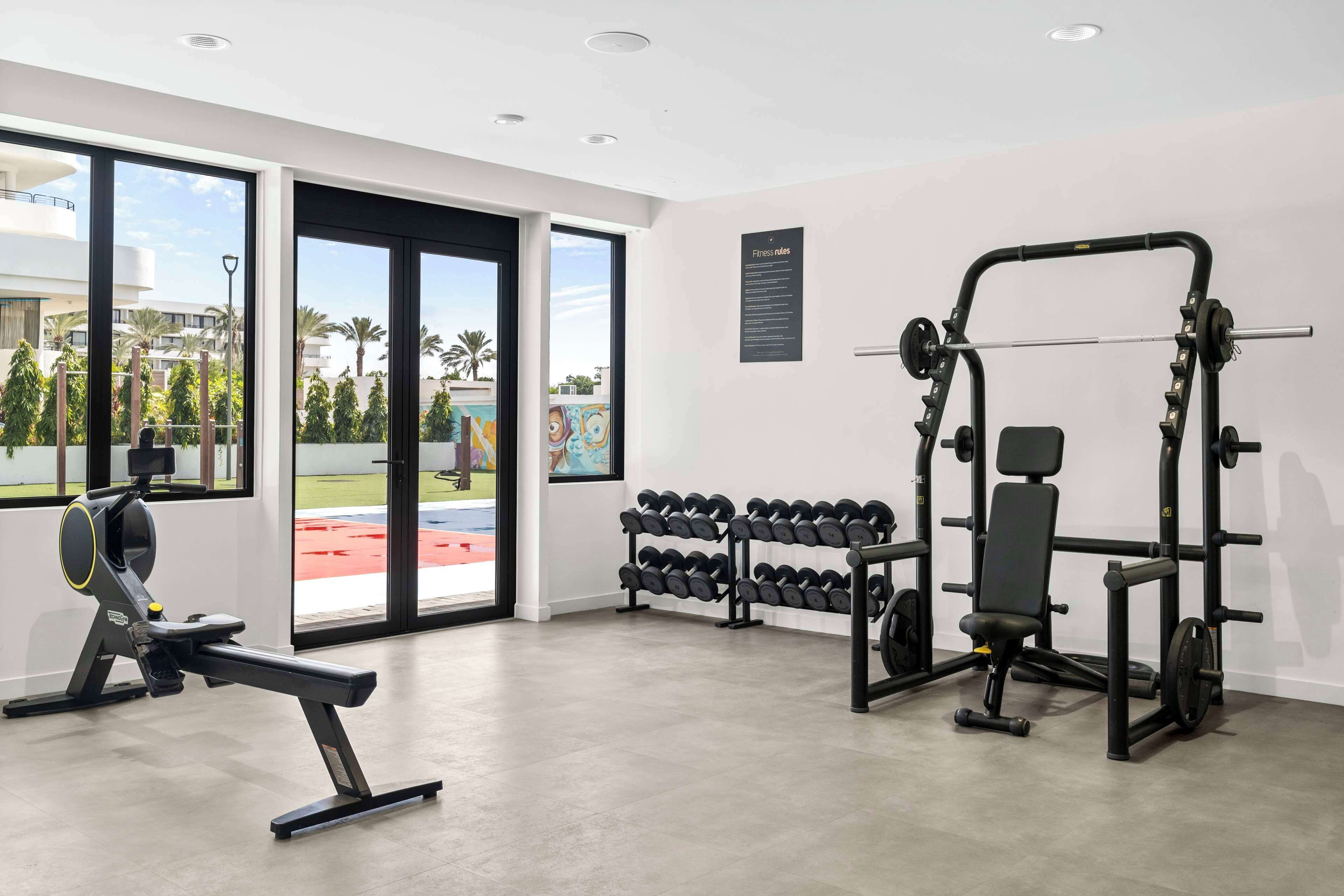 A gym with exercise equipment, large windows, and a view of palm trees.