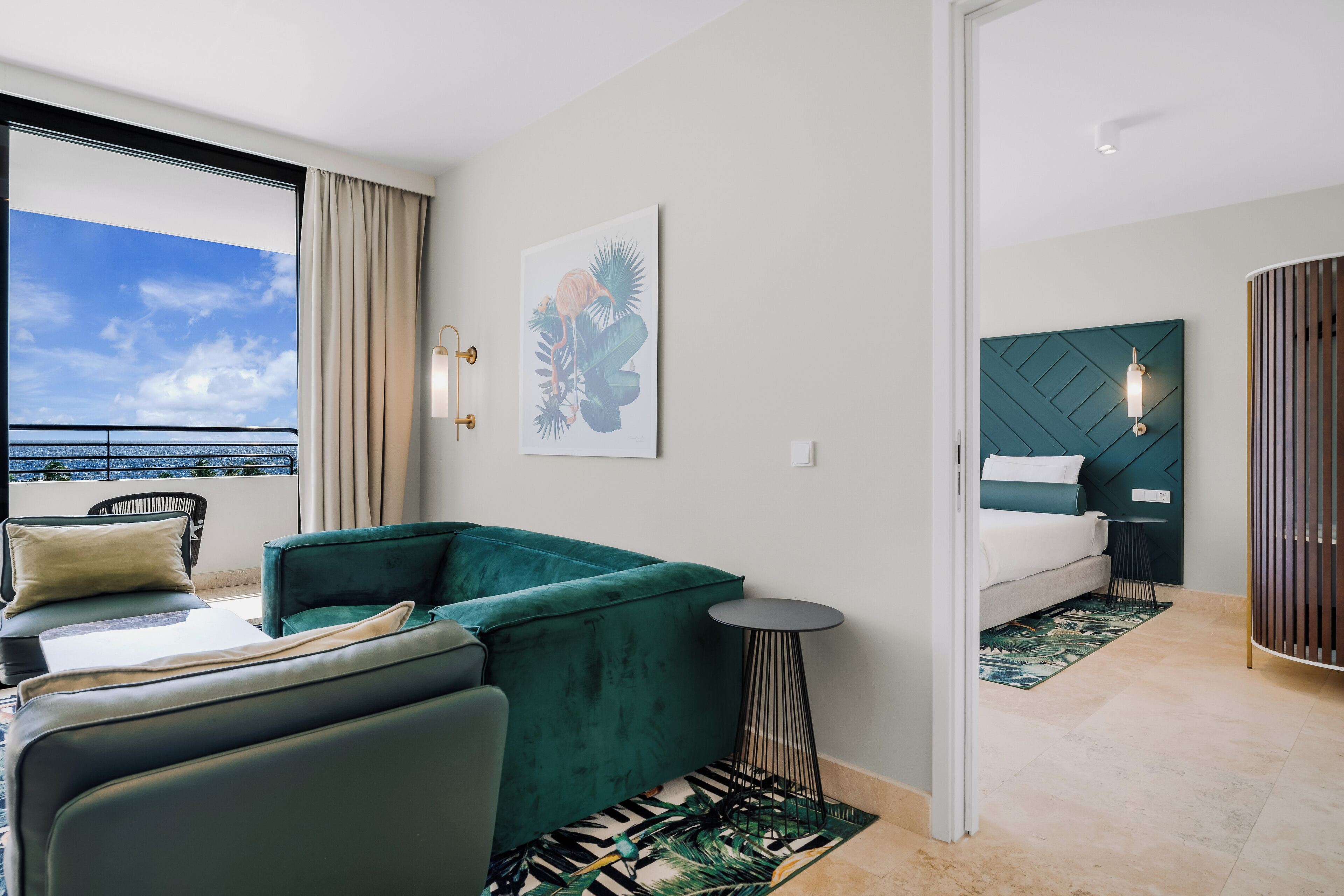 A modern hotel room with a balcony, a green sofa, a bed with a green headboard, and a tropical-themed artwork.