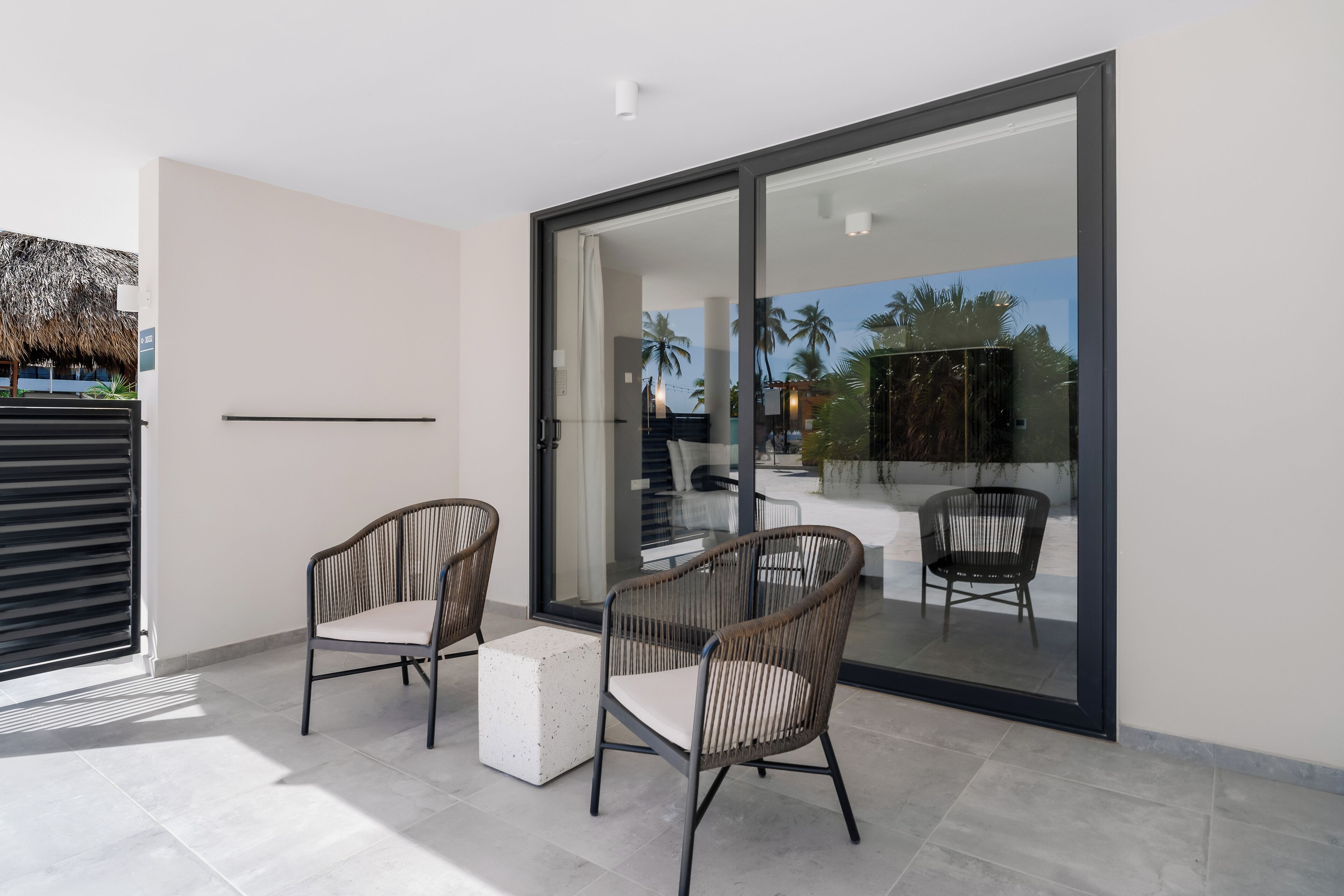 A modern patio area with two wicker chairs and a small white table, a sliding glass door leading to a pool area, and a view of palm trees and a building in the background.