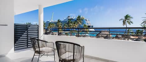 A balcony with two wicker chairs, a view of a cruise ship, palm trees, and a beach with thatched umbrellas.
