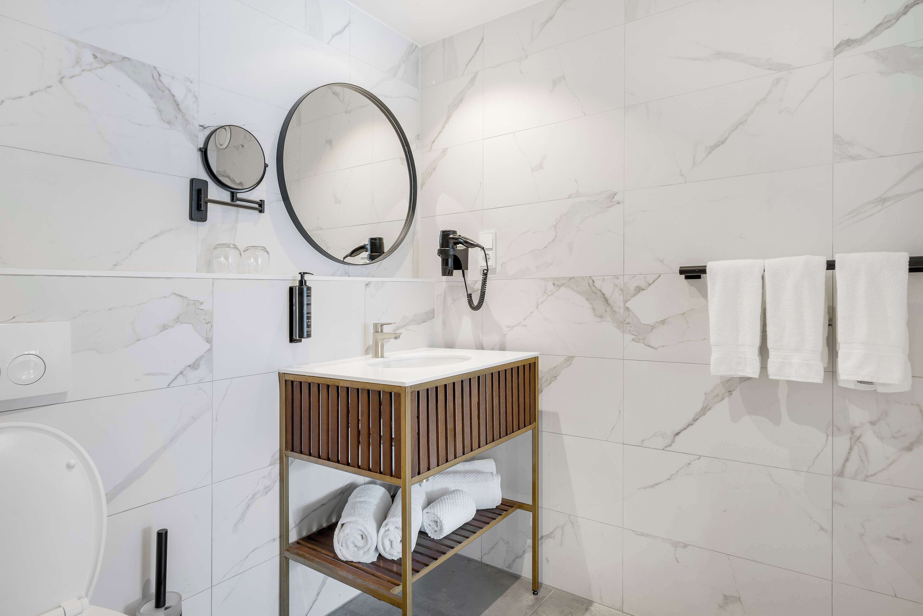 A modern bathroom with a marble-tiled wall, a wooden vanity with a sink, a mirror, a hairdryer, and towels hanging on a rack.