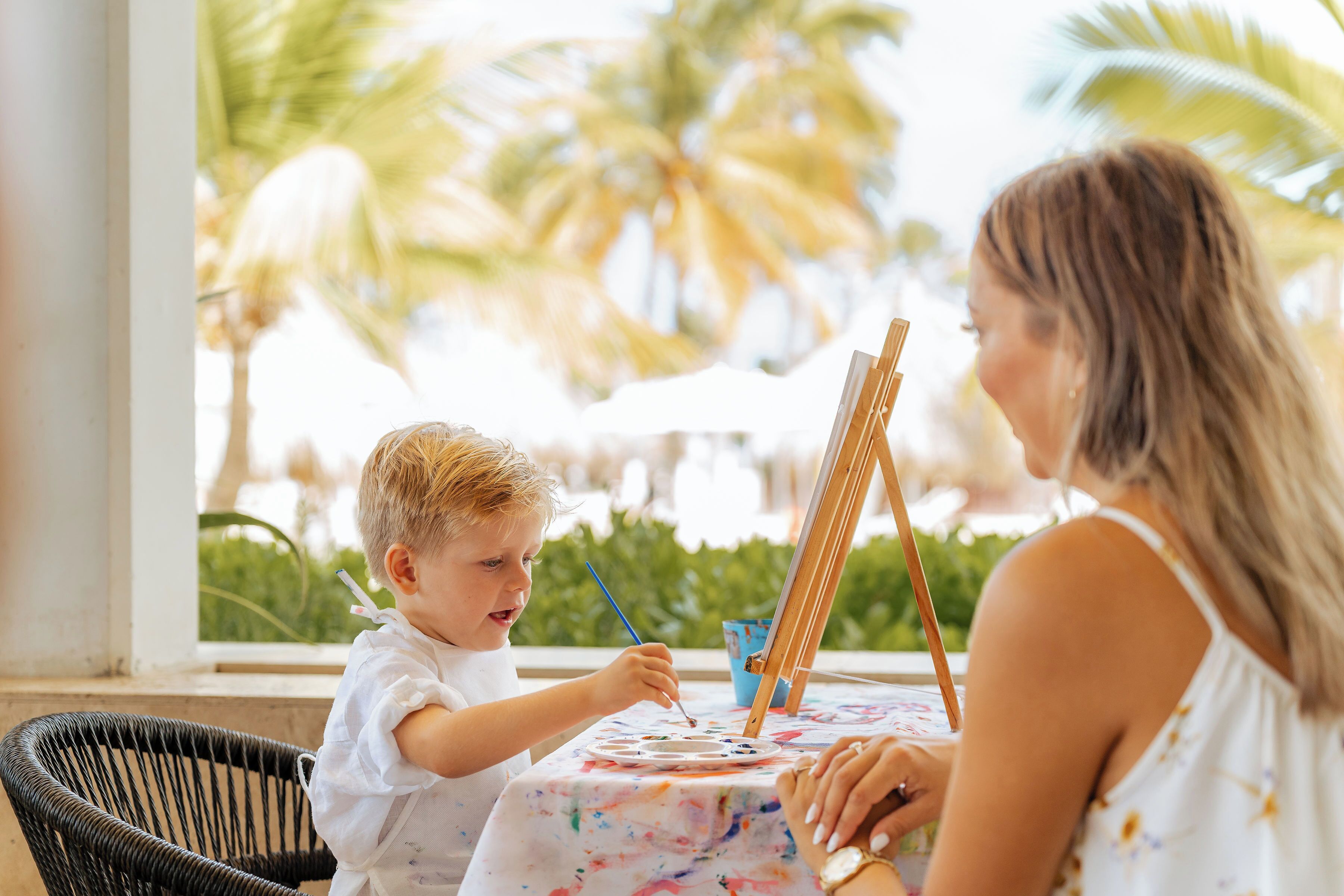 A child painting with an adult at a table with a view of palm trees.