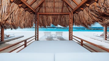 A beach cabana with a thatched roof, white cushioned seating, and a view of the ocean.