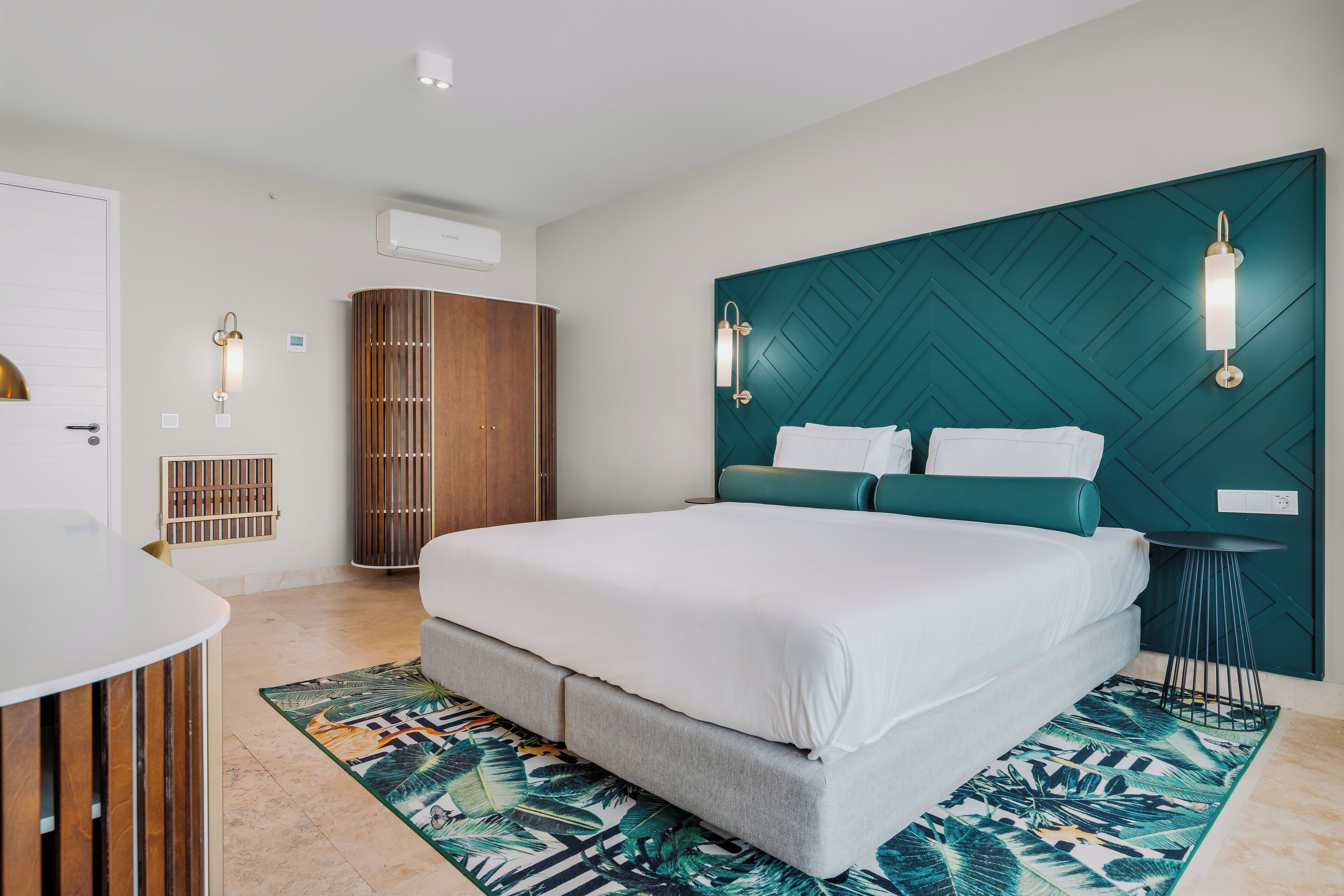A modern hotel room with a large bed, a wooden wardrobe, a small round table, and a decorative wall panel.
