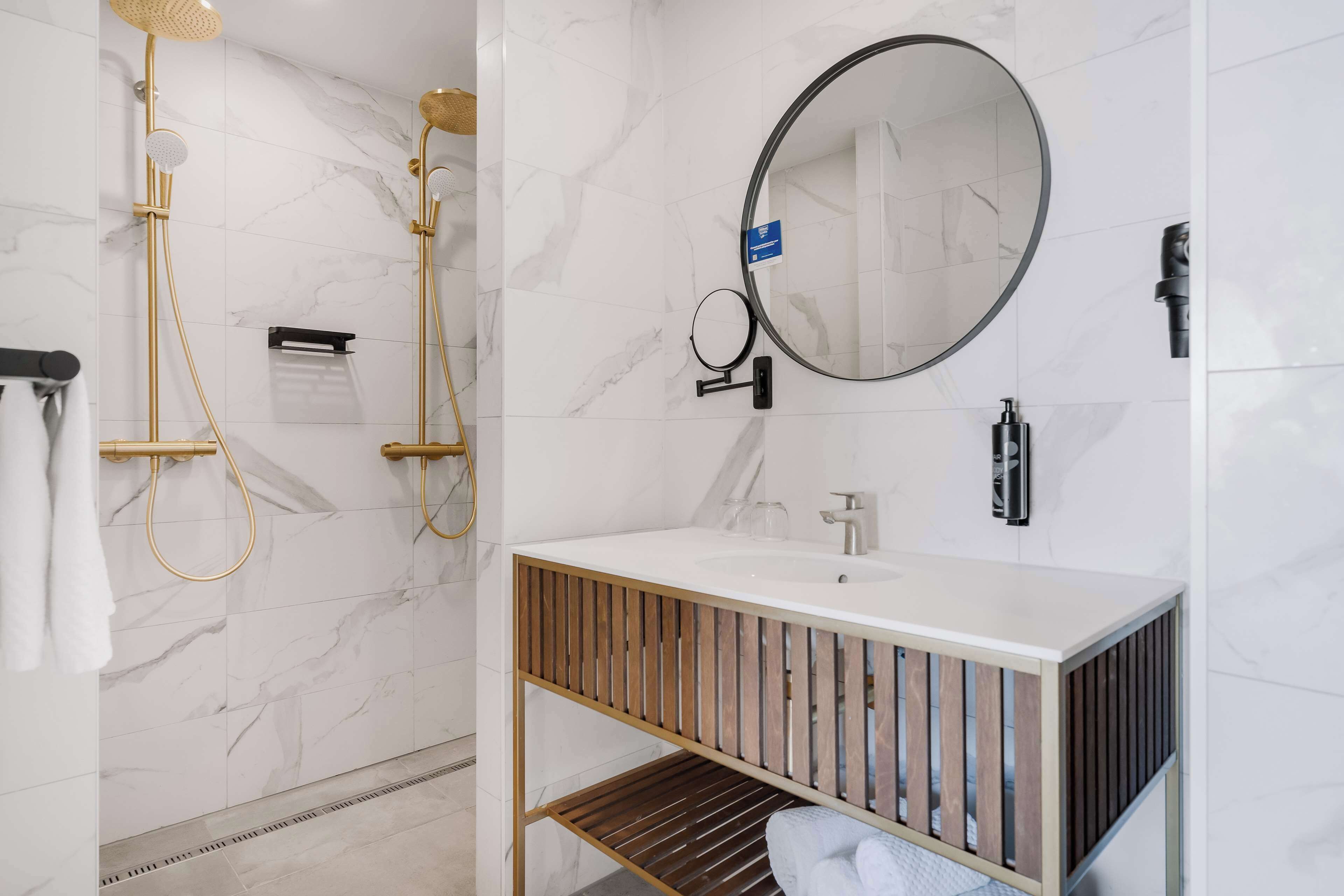 A modern bathroom with a white marble wall, a round mirror, a gold showerhead, and a wooden vanity.