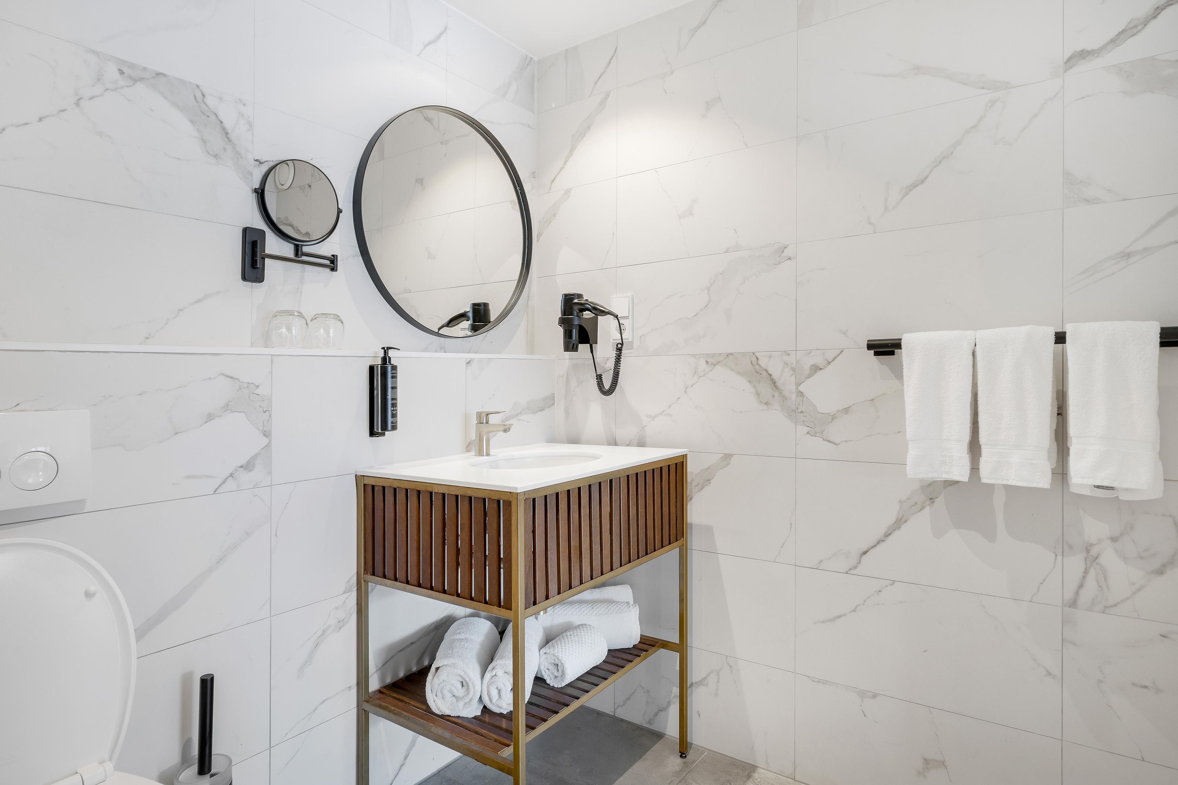 Un baño moderno con pared de mármol, un espejo redondo, un cabezal de ducha manual y un estante de madera con toallas.