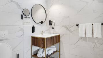 Un baño moderno con pared de mármol, un espejo redondo, un cabezal de ducha manual y un estante de madera con toallas.