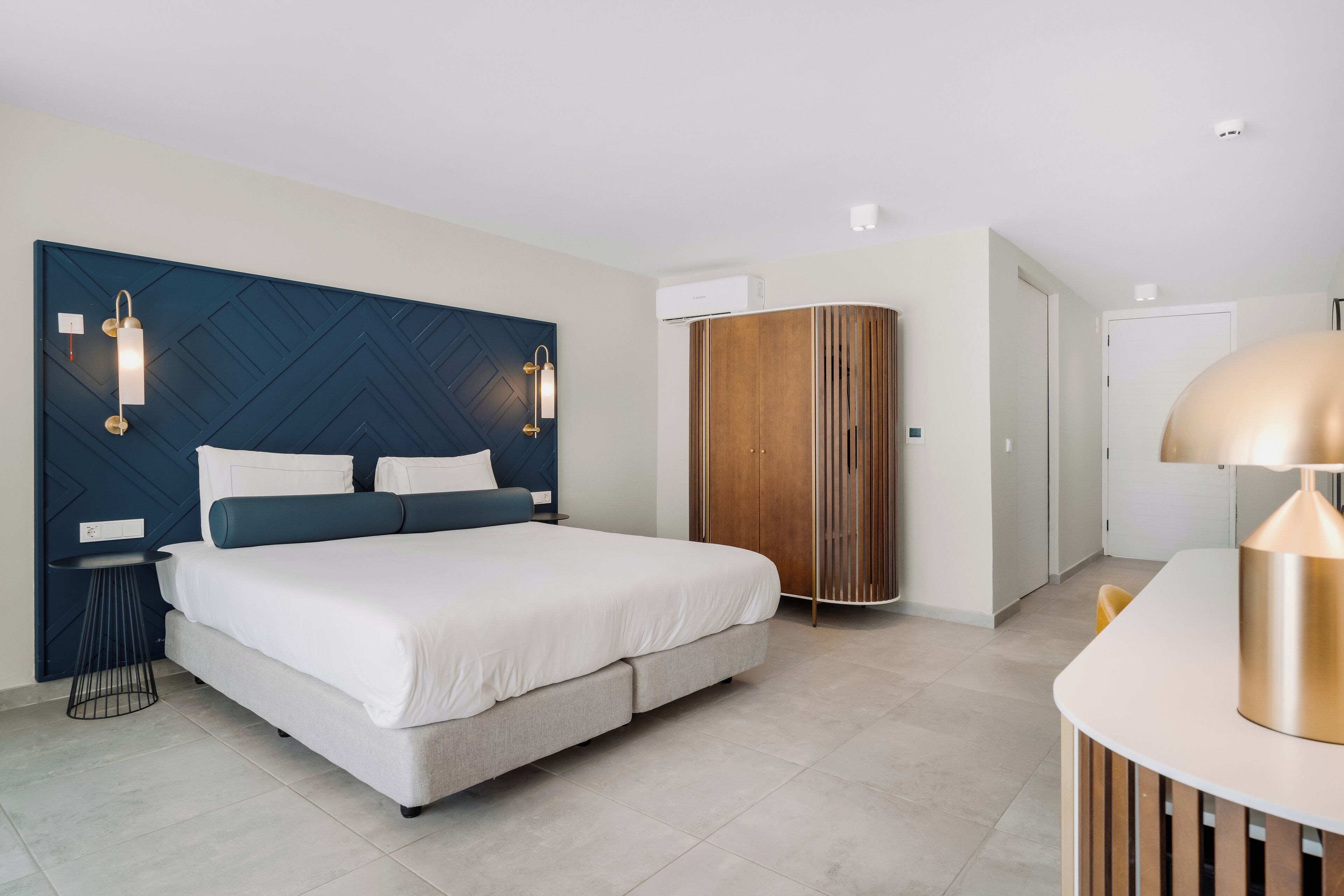 A modern hotel room with a large bed, a wardrobe, a desk, and a lamp.