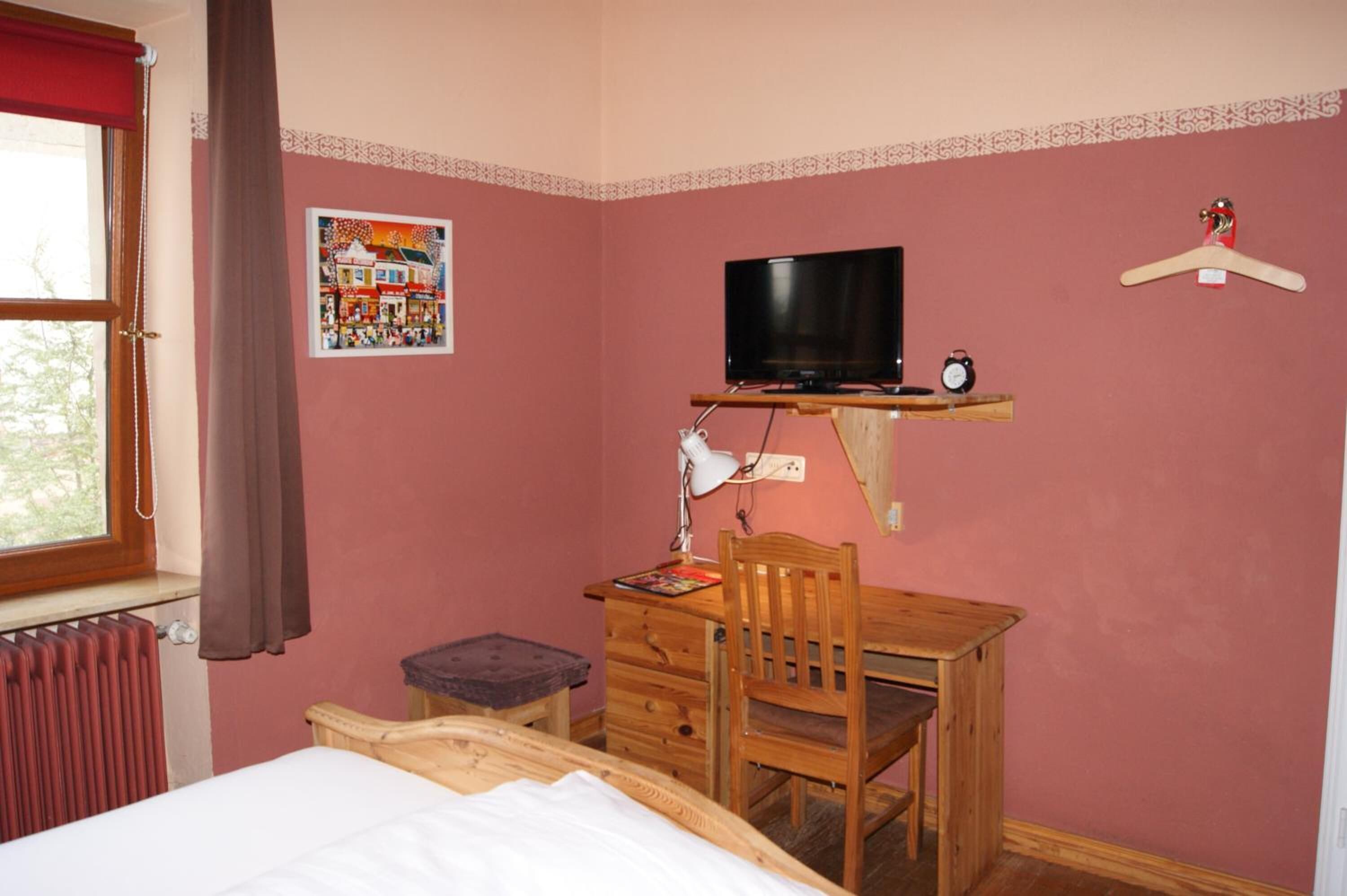 A bedroom with a bed, a wooden desk, a chair, a television, a radiator, and a window with curtains.