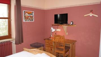 A bedroom with a bed, a wooden desk, a chair, a television, a radiator, and a window with curtains.