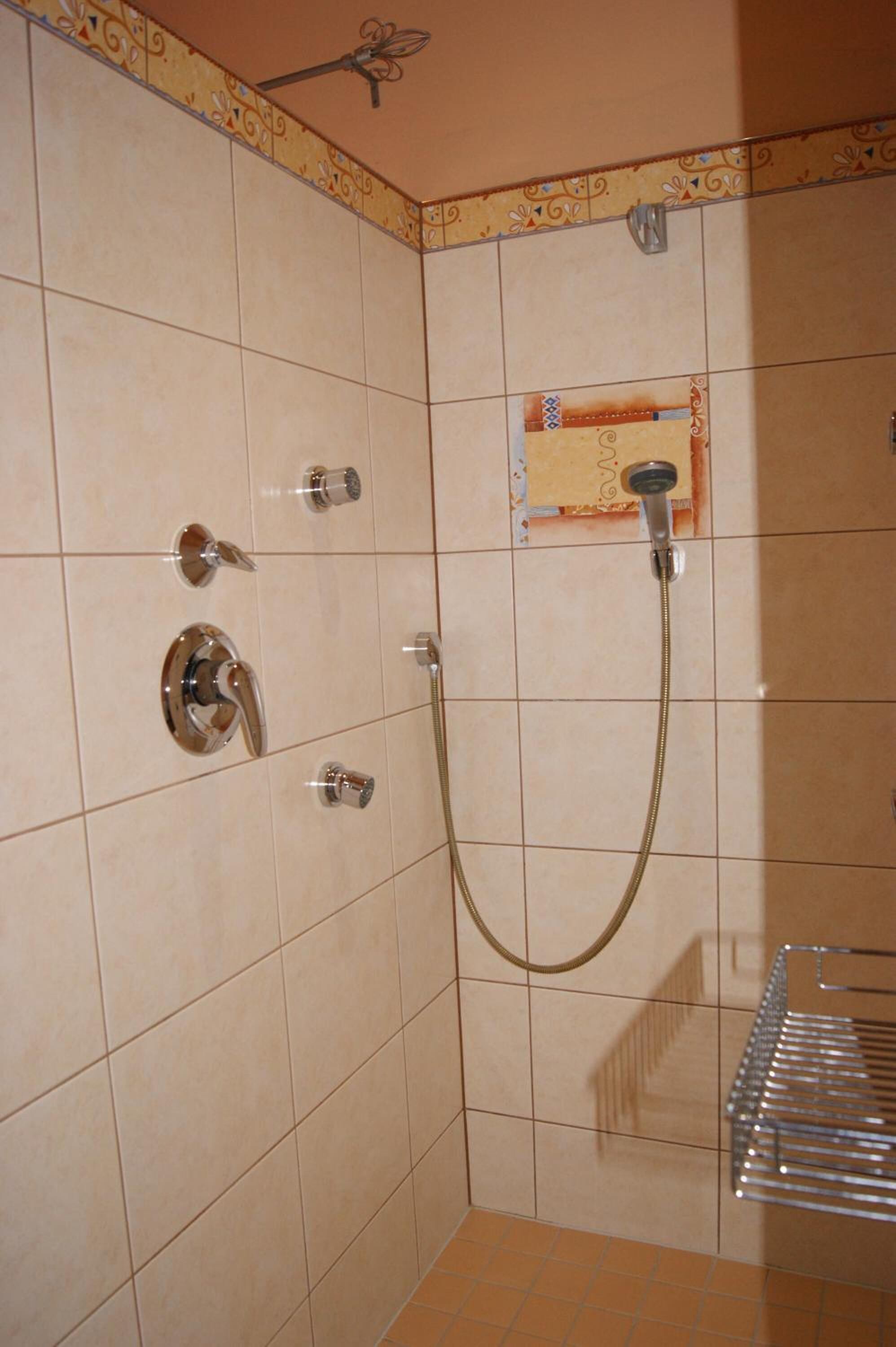 Ein Badezimmer mit beigen Fliesen, einer Dusche mit Duschhandstück und einem Metallregal.