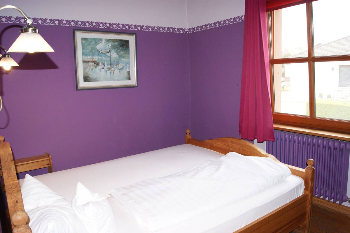 A single bed with a wooden frame and white bedding in a room with purple walls and a window with a red curtain.