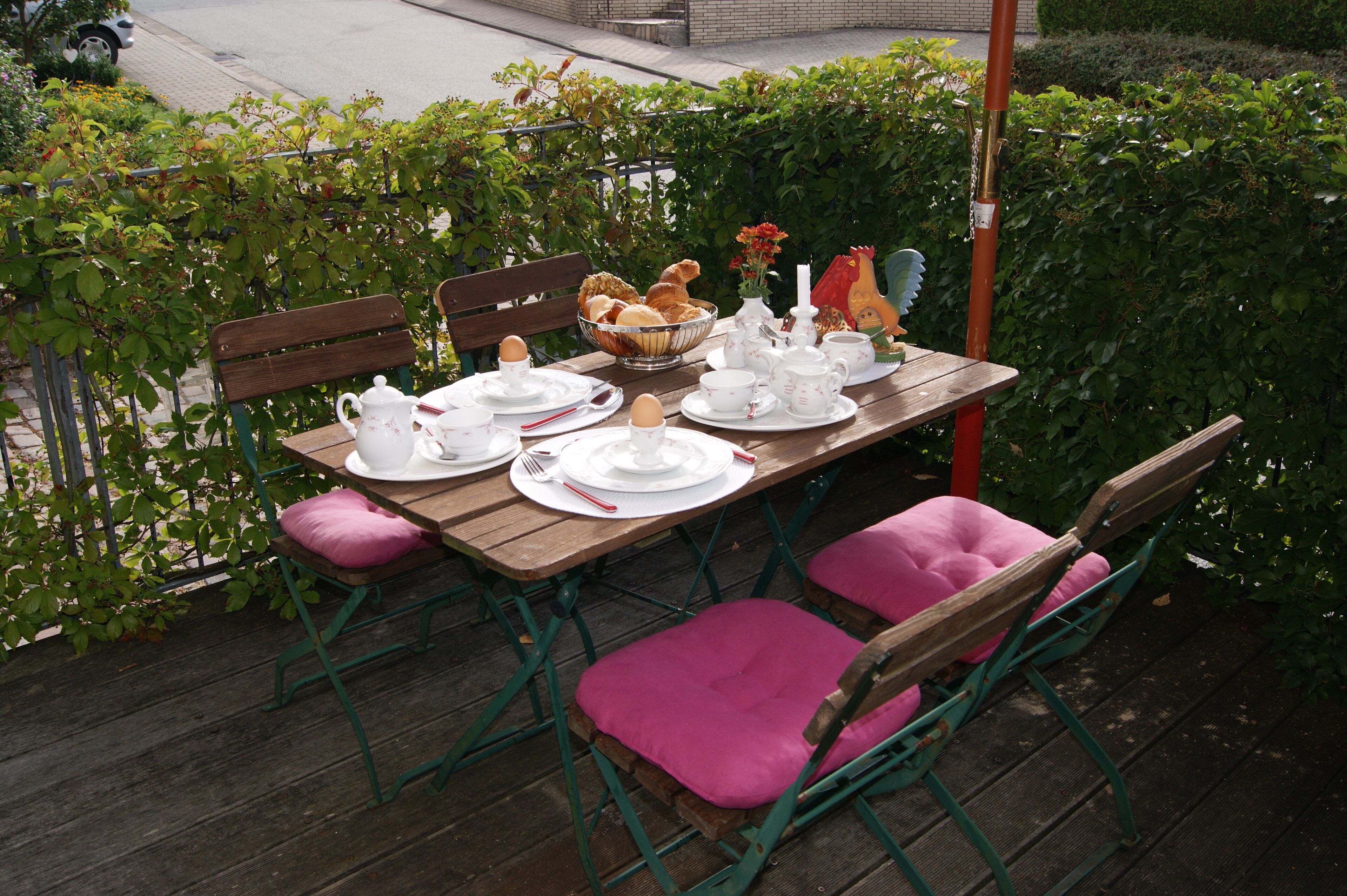 Un área de comedor al aire libre con una mesa de madera dispuesta para cuatro, rodeada de sillas de metal verde con cojines rosa.