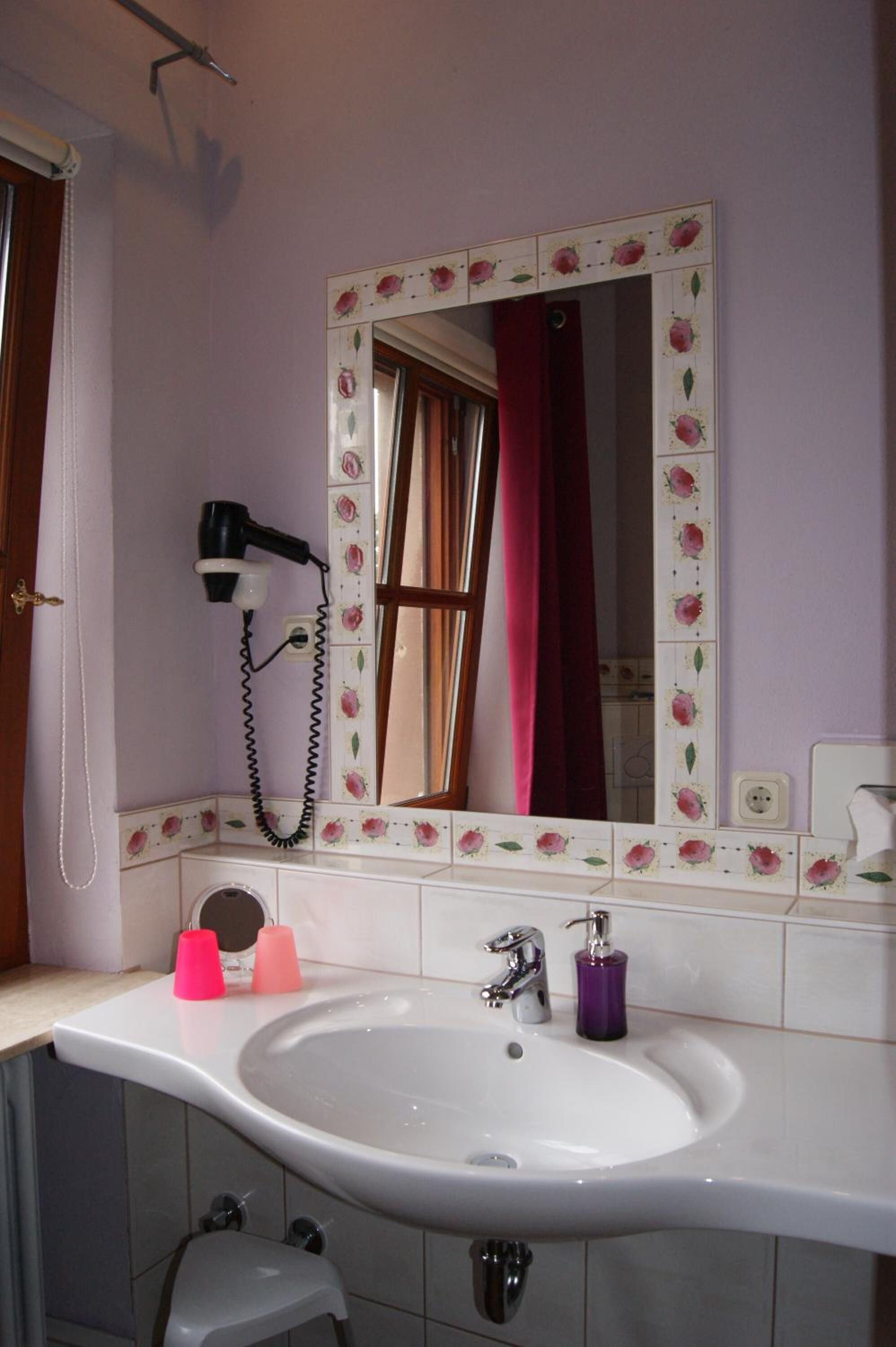 Ein Badezimmer mit einem weißen Waschbecken, einem Spiegel mit floralem Dekor, einem Fön und einem roten Duschvorhang.