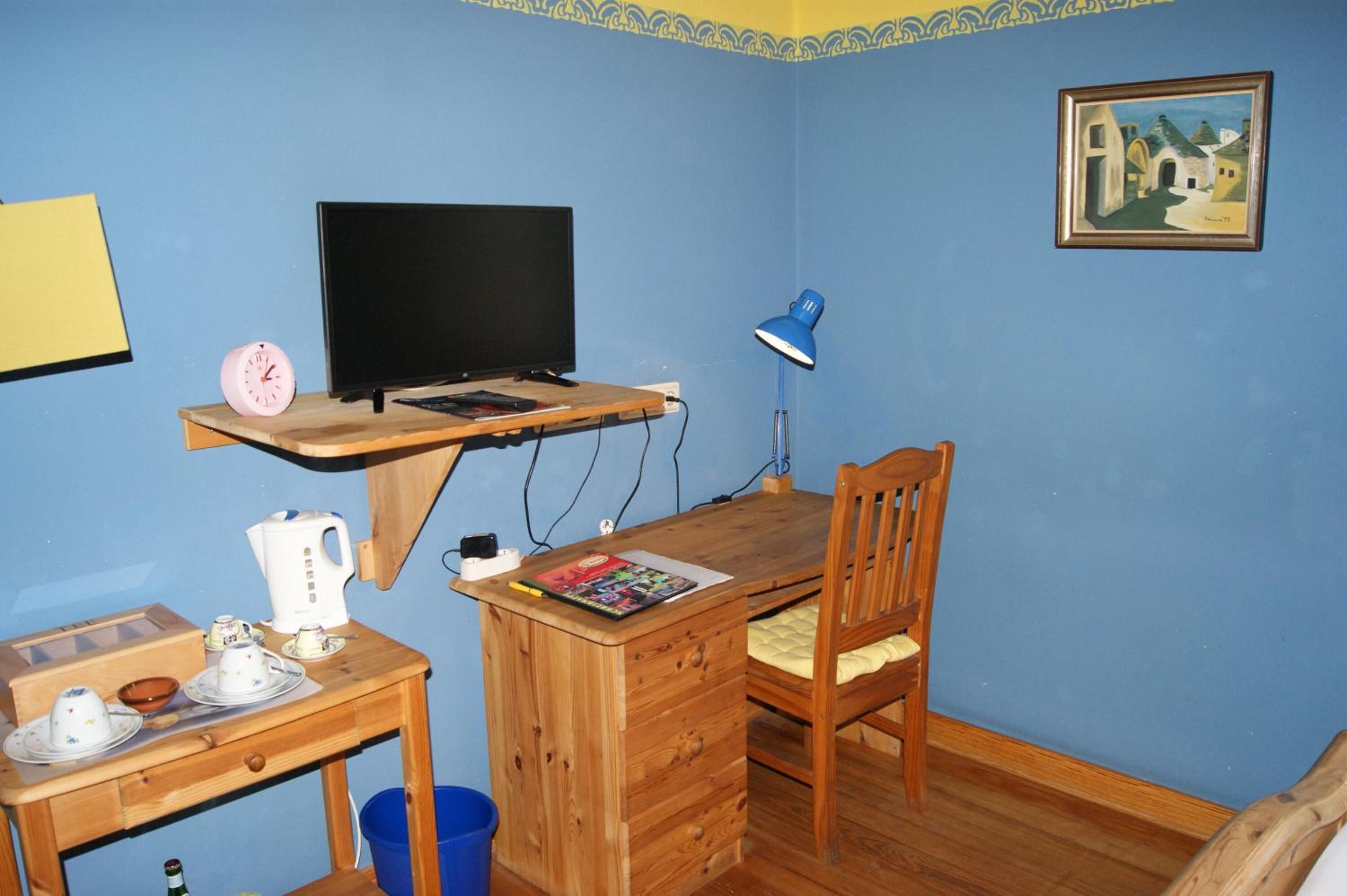 Ein kleines Zimmer mit einem Holztisch, einem Stuhl, einem Fernseher, einer Lampe und einem Bild an der Wand.