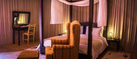 A four-poster canopy bed with a wooden chair and ottoman, a desk with a chair, and a nightstand with a lamp.