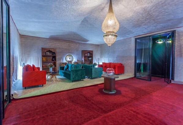 Una sala de estar moderna con sofás rojos, un sofá verde y una mesa de centro. Hay una araña colgando del techo y una puerta de vidrio que da al exterior.