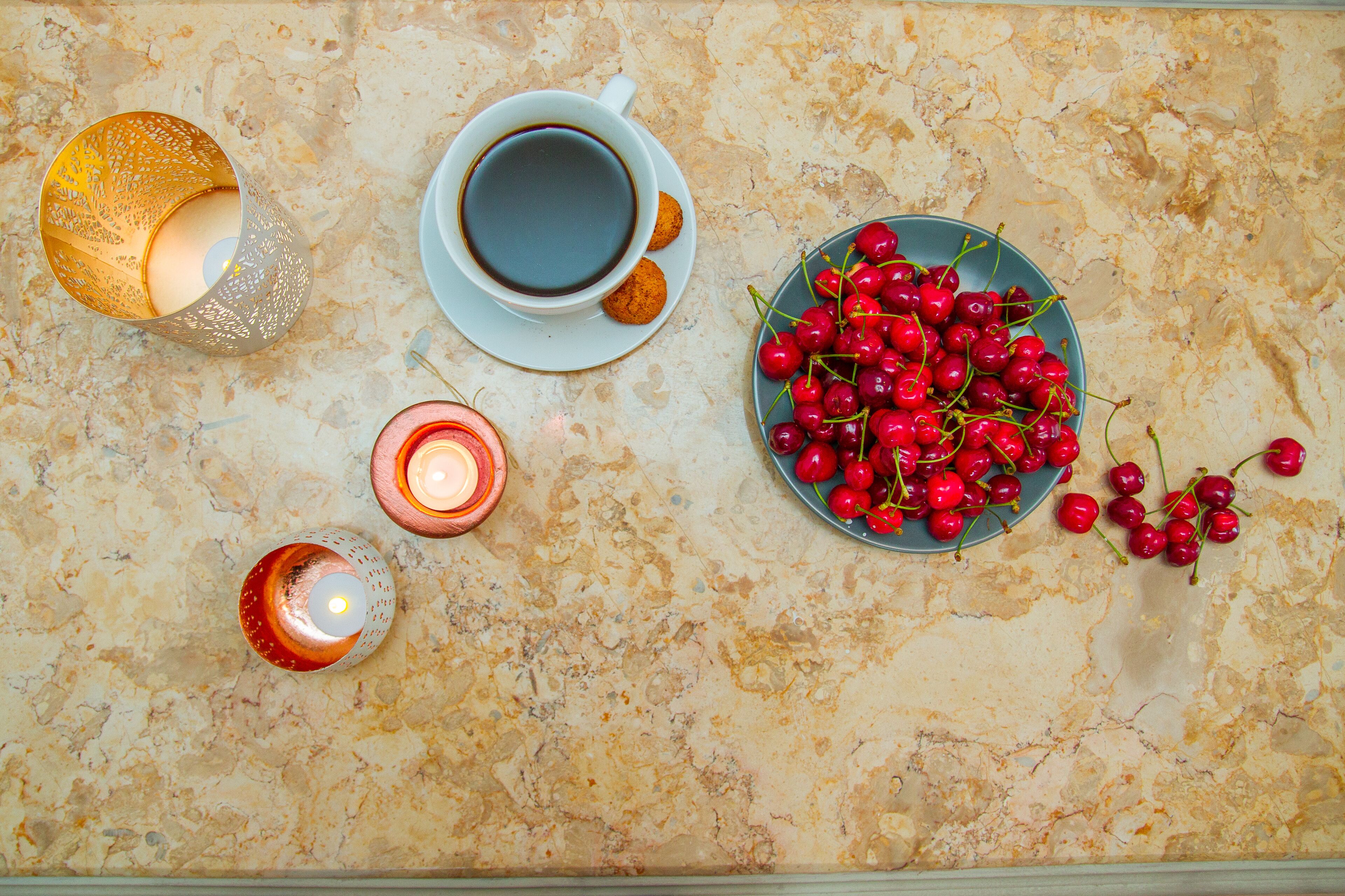 Une table avec une tasse de café, une assiette de biscuits, un bol de cerises et deux bougies.
