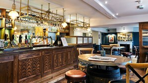 A bar area with wooden paneling, a counter with multiple taps, hanging lights, and a round table with chairs. - The Wellington Arms (Hook)