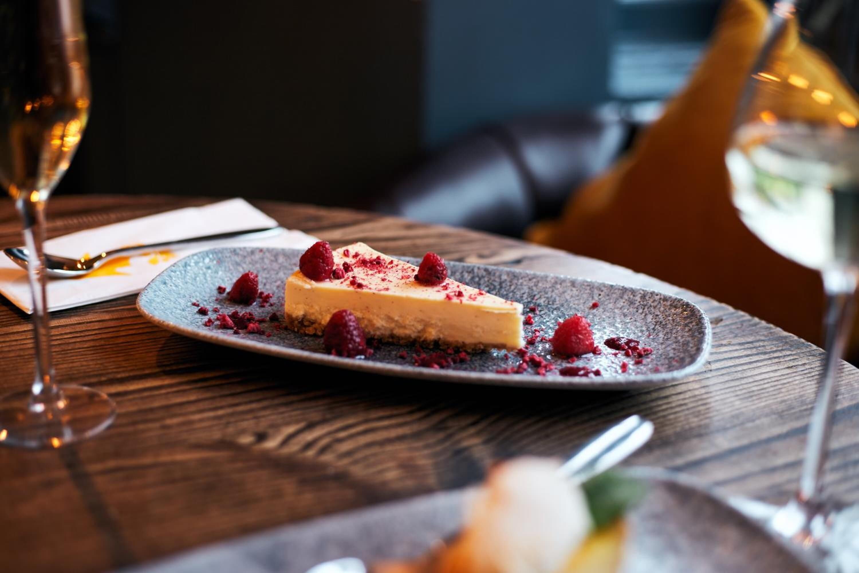 Un plato de postre con un pastel de queso, frambuesas y una copa de vino blanco sobre una mesa de madera.