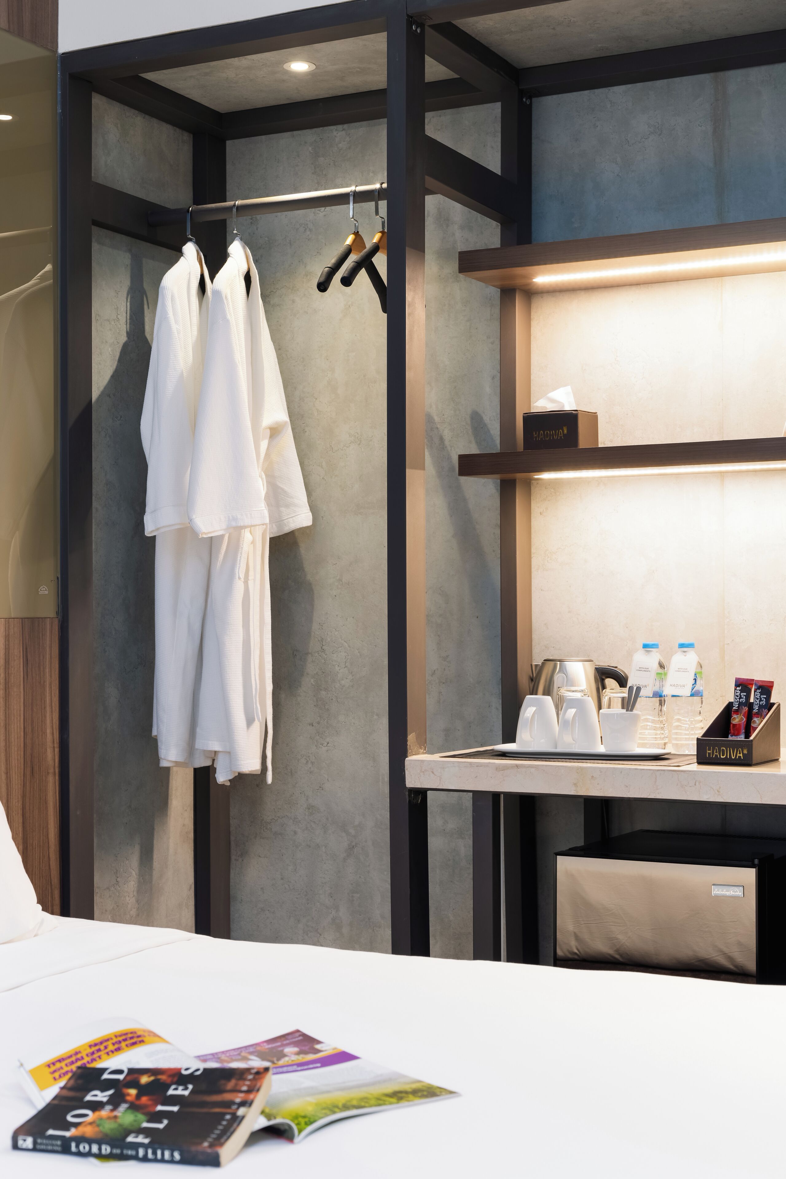 A hotel room with a bed, a robe rack with white robes, a small table with a coffee maker, cups, and a box of tissues.