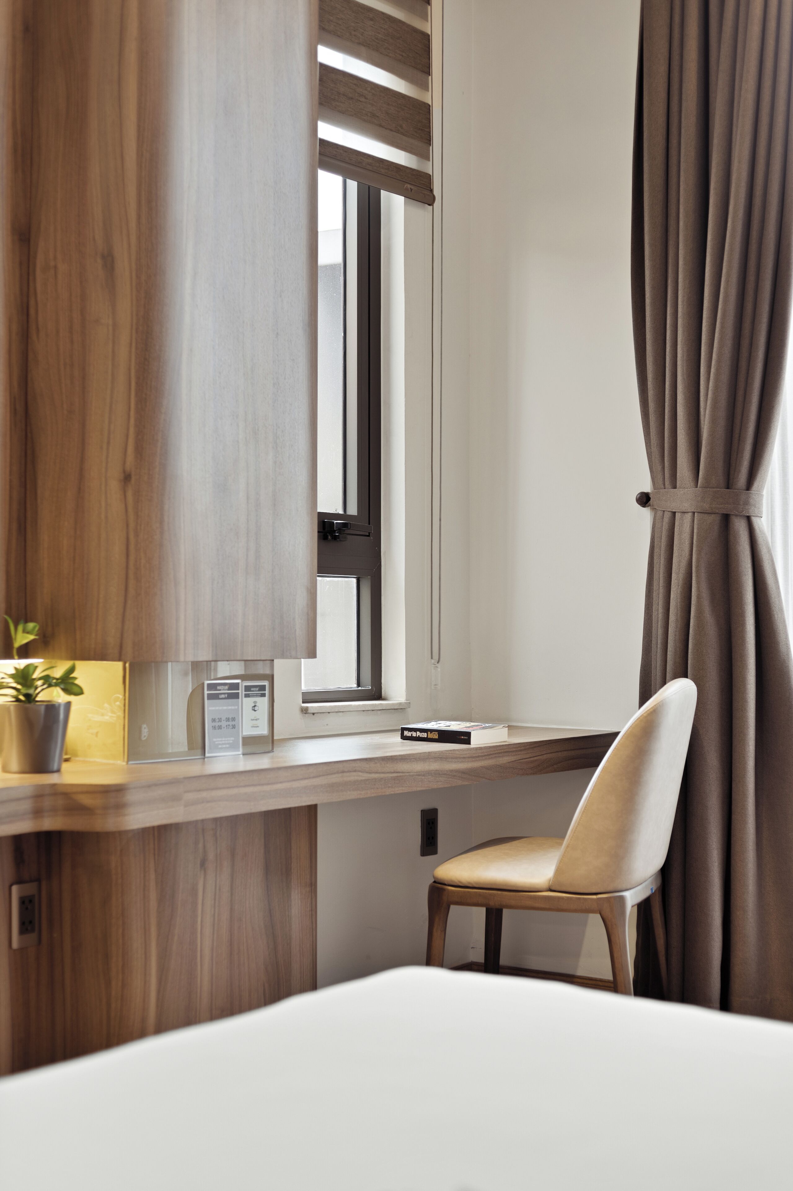 A hotel room with a wooden desk, a chair, a potted plant, and a window with curtains.