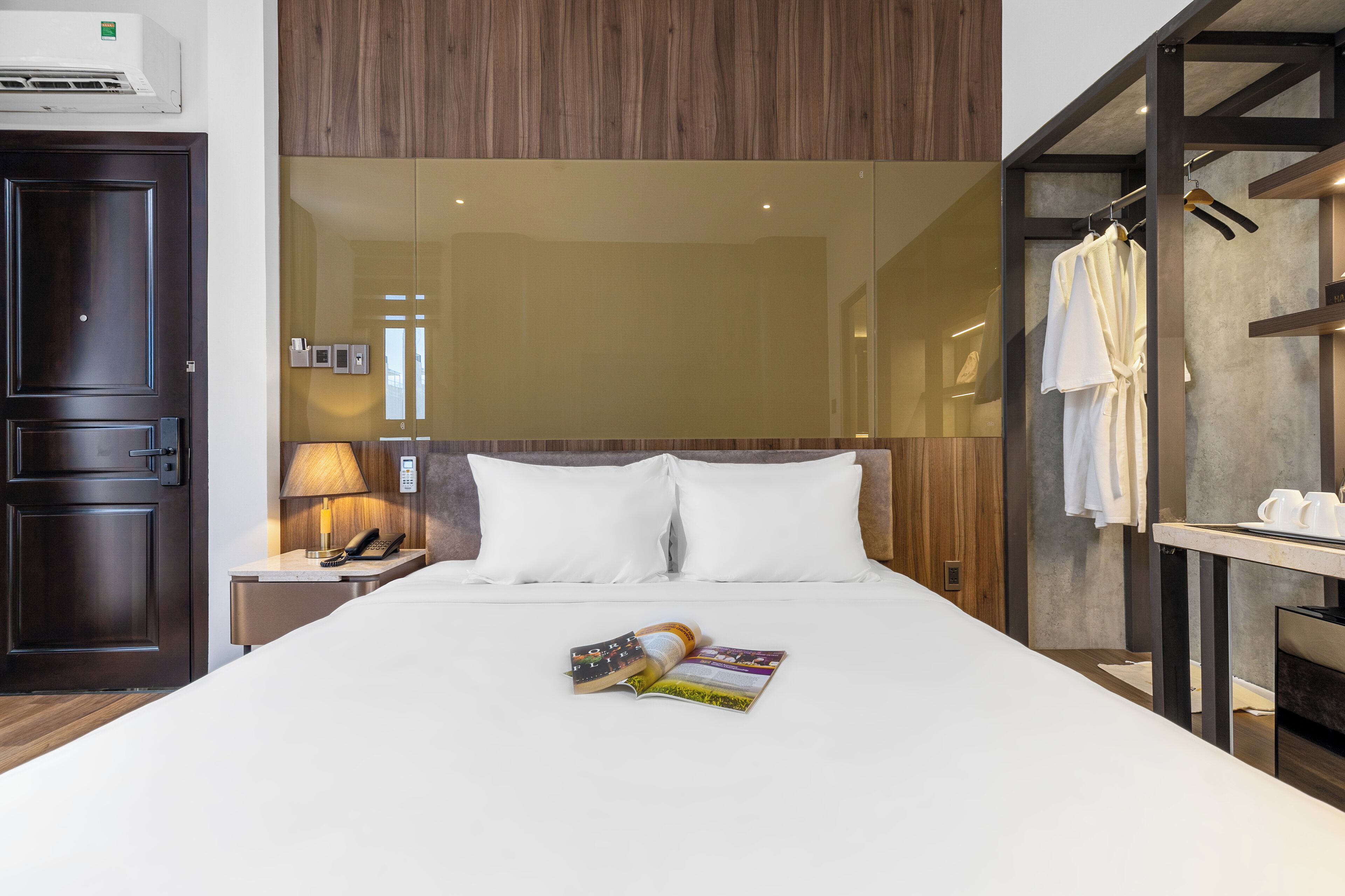 A hotel room with a bed, a nightstand with a lamp, a phone, and a magazine. There is a wooden door and a wardrobe with clothes hanging.