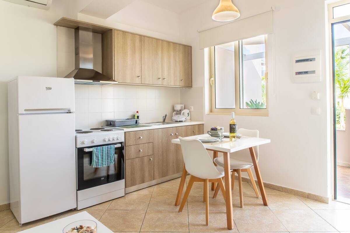 A modern kitchen with a white refrigerator, stove, and wooden cabinets. There is a dining table with chairs and a window with a view of greenery outside.