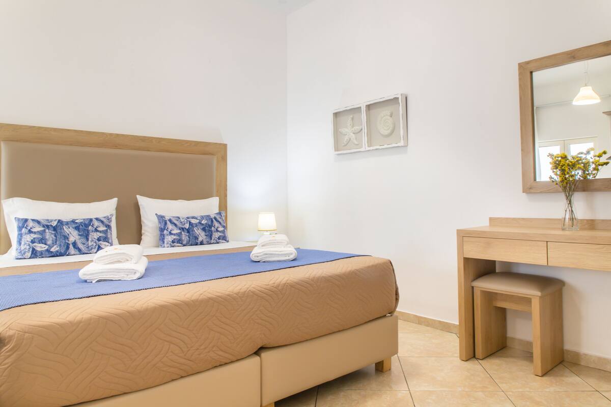 A hotel room with a bed, a bedside table, a mirror, and a small wooden stool.
