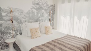 Une chambre à coucher avec un lit, du linge de lit blanc, un plaid à motifs et un mur orné d’une fresque représentant un arbre.