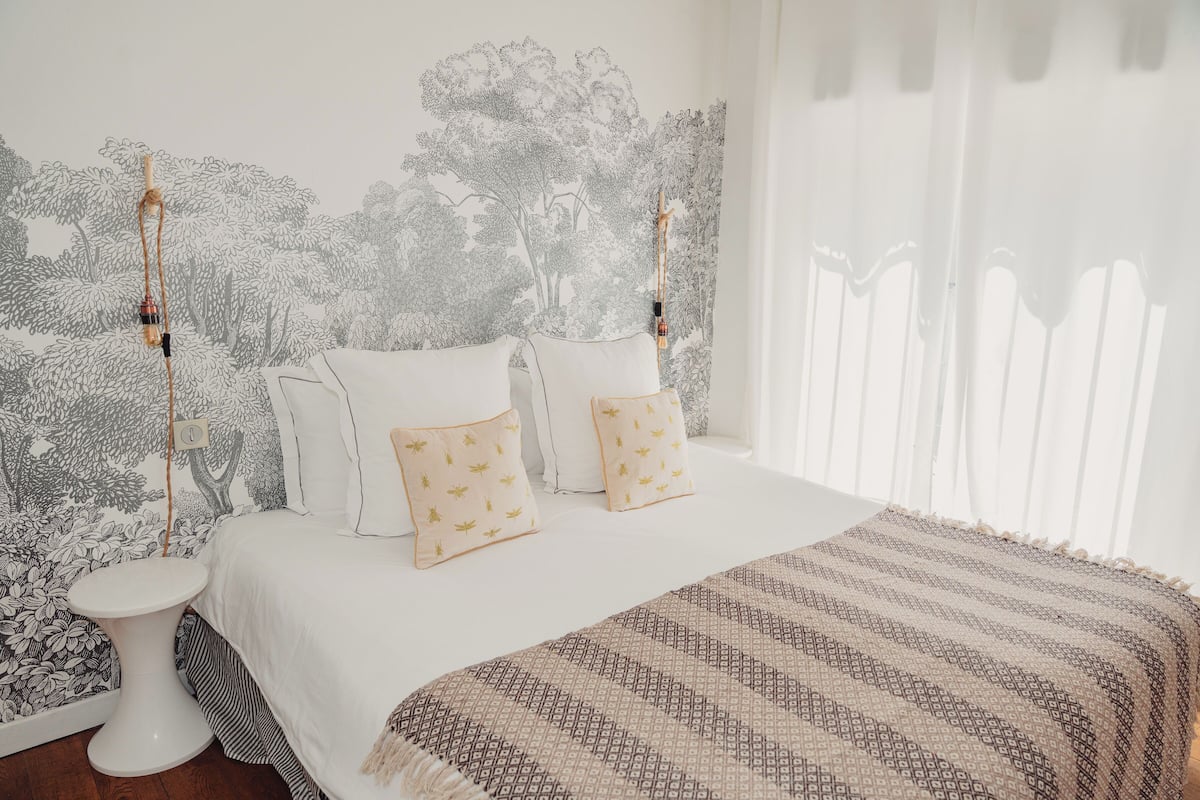 Un dormitorio con cama, ropa de cama blanca, manta estampada y una pared con un mural de un árbol.
