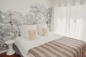 Un dormitorio con cama, ropa de cama blanca, manta estampada y una pared con un mural de un árbol.