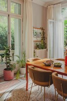 Comedor con mesa de madera y sillas de mimbre, rodeado de plantas en macetas y vistas al verdor exterior.