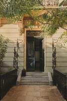 Entrada de un edificio con una escalera que conduce a una puerta, rodeada de rejas decorativas y plantas.