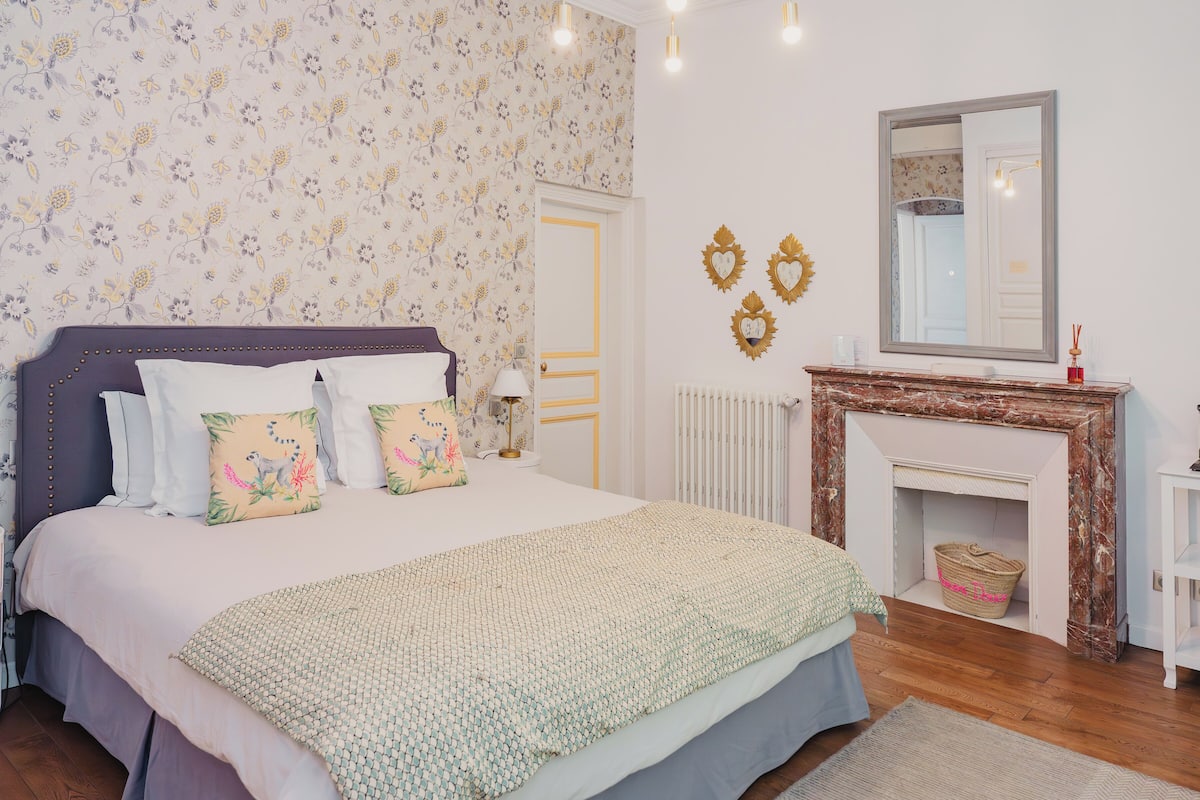 Un dormitorio con una cama grande, papel tapiz floral, chimenea y un espejo en la pared.