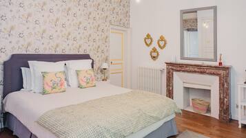 Une chambre avec un grand lit, un papier peint à motifs floraux, une cheminée et un miroir au mur.