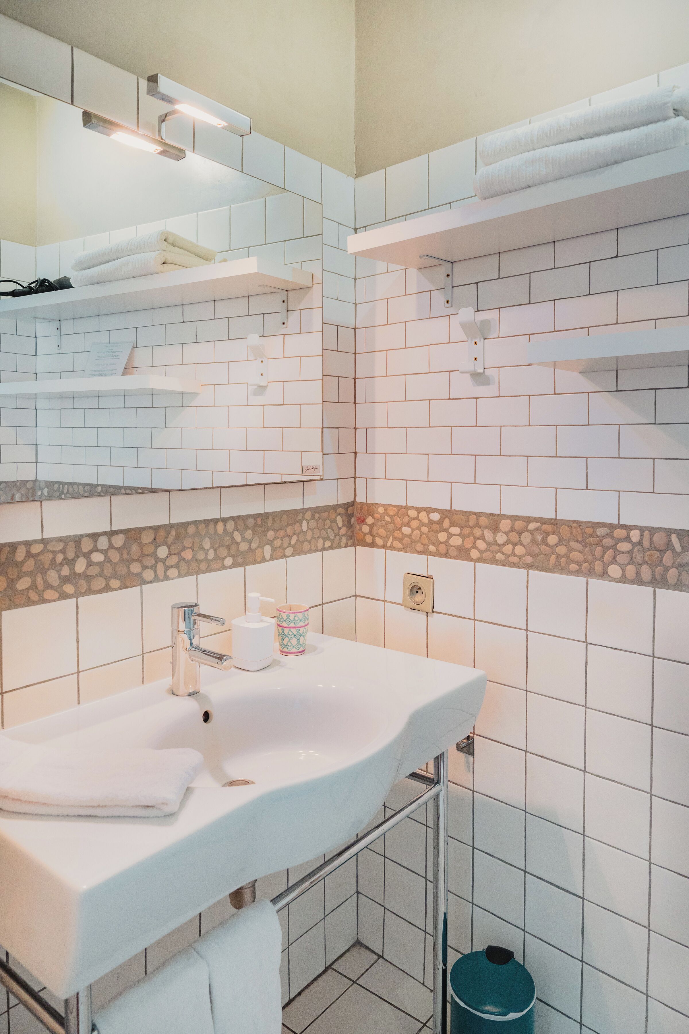 Een badkamer met witte betegelde wanden, een witte wasbak en een spiegel erboven. Er zijn twee handdoekrekken met handdoeken erop.