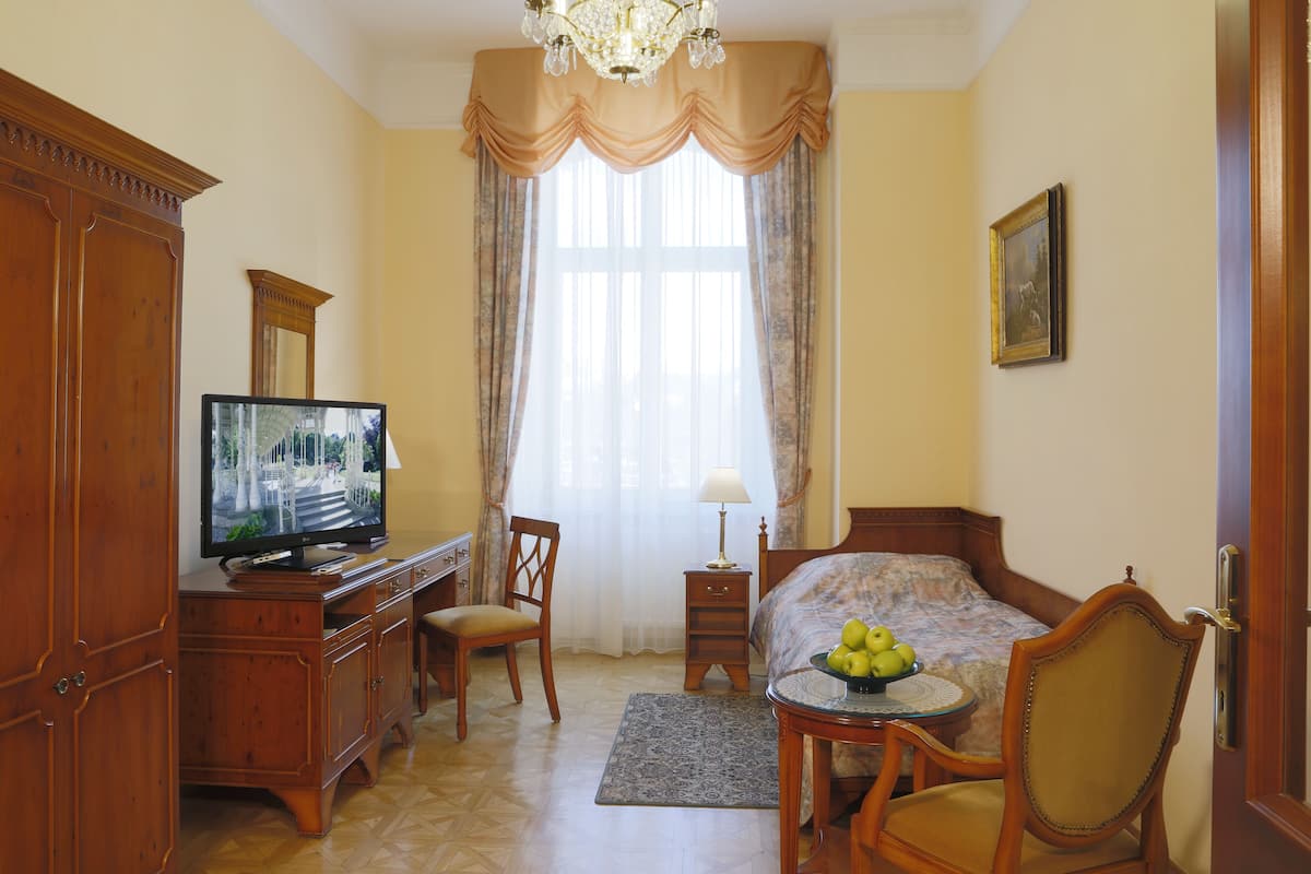 Une chambre d’hôtel avec une commode en bois, un téléviseur, un lit recouvert d’une couvre-lit à motifs floraux, une chaise, une petite table avec des fruits et une fenêtre avec des rideaux.