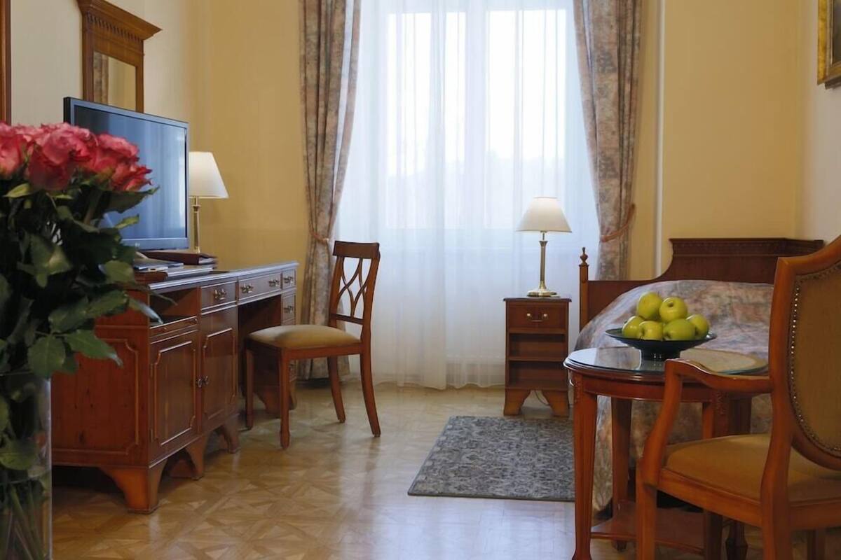Une chambre d’hôtel avec un bureau en bois, une chaise, un lit, une télévision, une lampe, un vase de fleurs et une fenêtre avec des rideaux.
