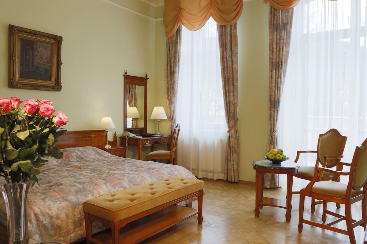 Une chambre d’hôtel avec un lit, un bureau, deux chaises, une petite table avec des fruits, un vase avec des fleurs et un tableau au mur.